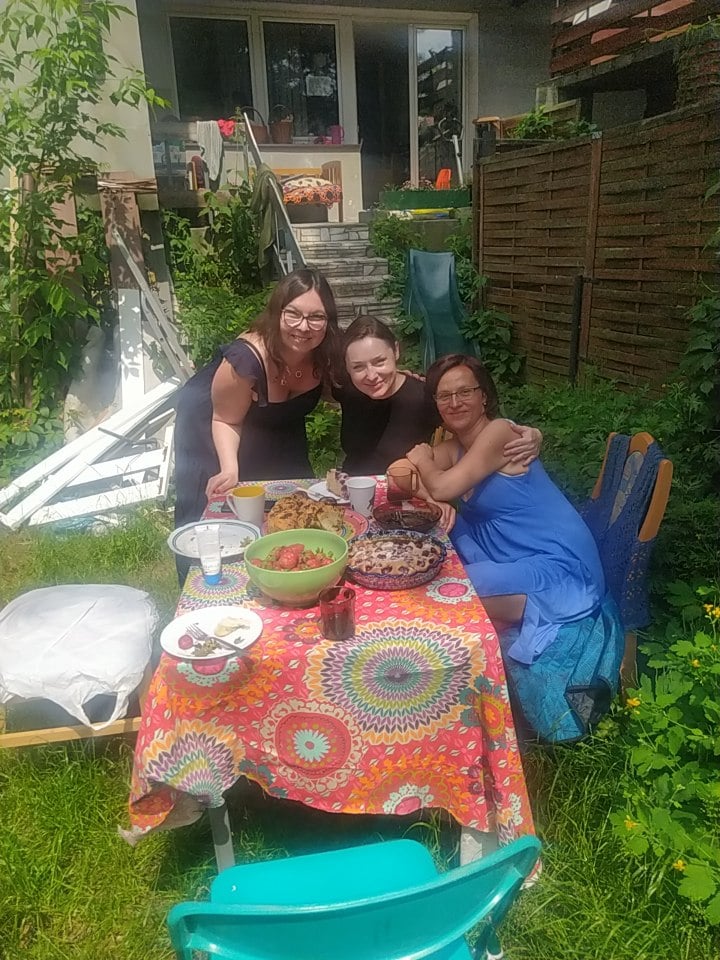 trzy uśmiechnięte kobiety siedzą przy stole, w ogrodznie, przy drewnianym domu