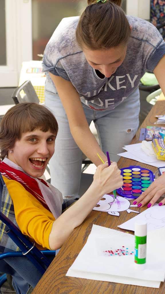 Uśmiechnięta osoba z niepełnosprawnością maluje z pomocą kobiety
