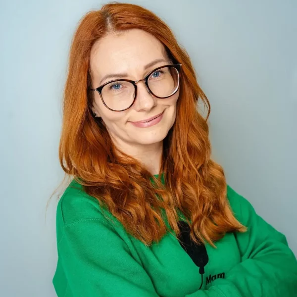 Kobieta w okularach z długimi, rudymi włosami, ubrana w czarną marynarkę.