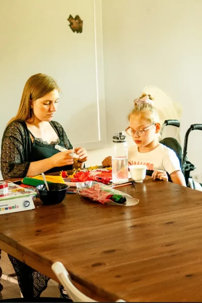 Wolontariuszka i dziewczynka wspólnie wykonują prace plastyczne przy drewnianym stole